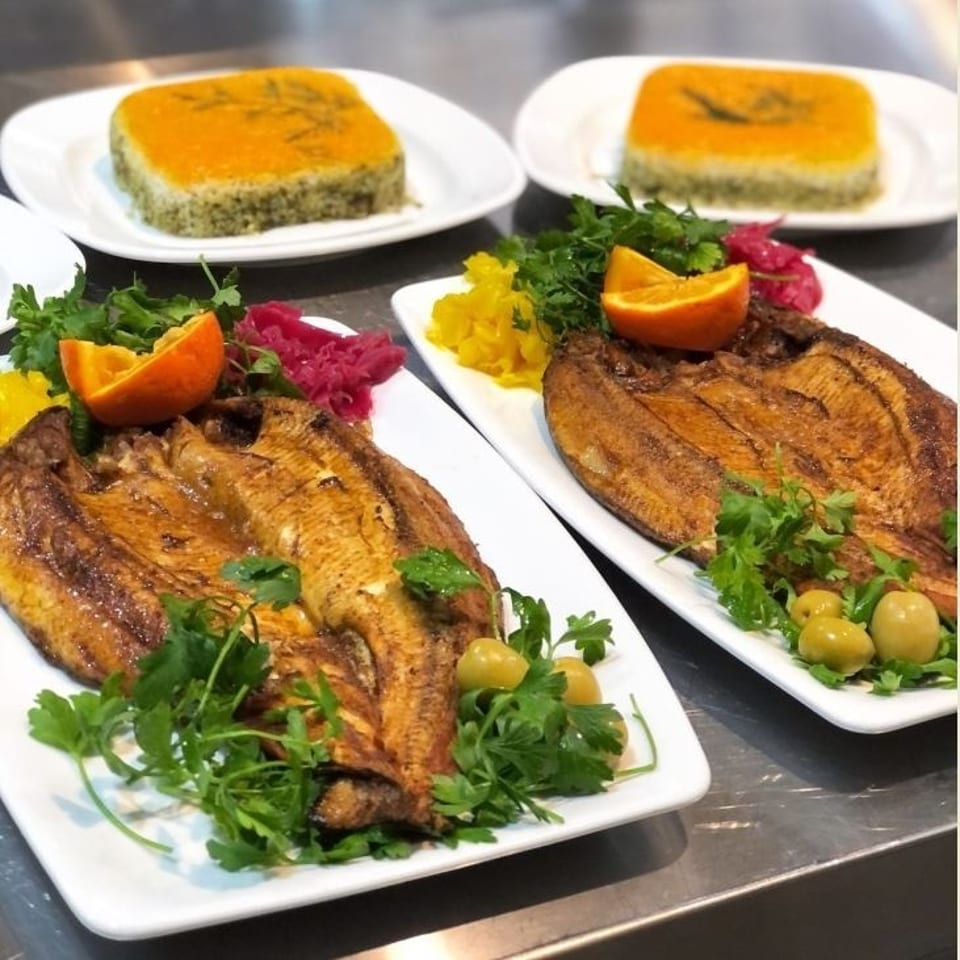 Two plates of grilled fish garnished with parsley, orange wedges, olives, pickled vegetables, and square golden desserts in the background.