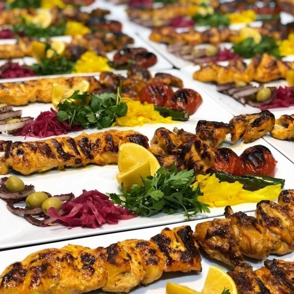 Rows of grilled chicken skewers and vegetables arranged on white platters with lemon wedges, parsley, olives, and pickled onions.