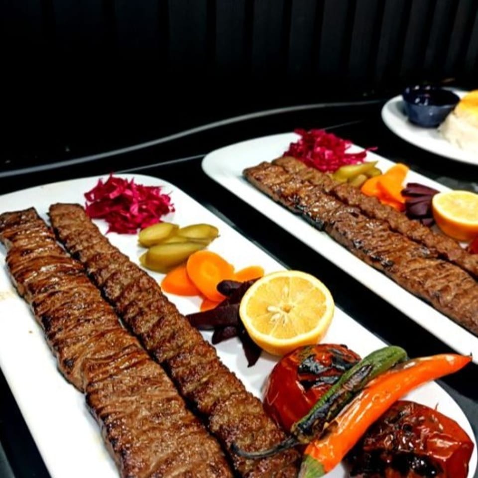 Grilled kebab platters with roasted vegetables, pickles, red cabbage, and lemon halves on white plates.